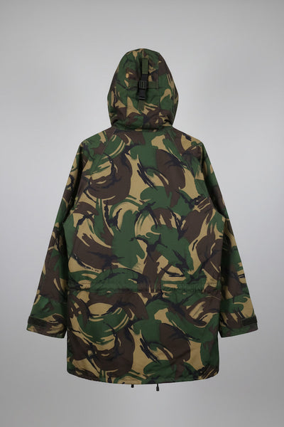 A hooded camouflage jacket shown from the back, featuring an all-over abstract leaf-like pattern in green, brown, and tan, photographed against a neutral gray background.