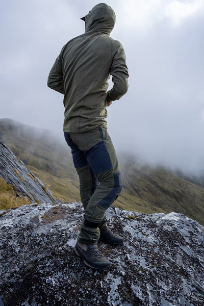 A person standing on a rocky mountain ridge, viewed from behind, wearing an olive green hooded jacket, dark blue and gray technical pants, and hiking boots, with misty hills in the background.
