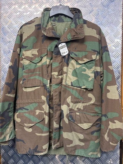 A woodland camouflage military-style jacket with four front flap pockets, hanging on a black hanger against a silver diamond plate metal background. A white hangtag is attached near the collar.