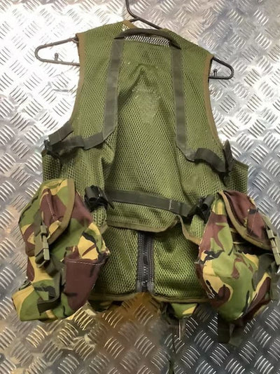 An olive green tactical vest with camouflage utility pouches on the sides, hanging on a metal hanger over a diamond plate metal background.