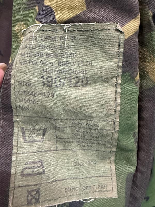 Close-up of a camouflage garment interior label showing printed text including 'Height/Chest 190/120' and care symbols, with a hand holding the fabric edge.