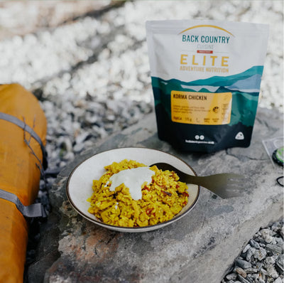 Campfire meal with a bowl of food and a 'Back Country Elite' package on a rocky surface.