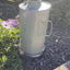 Metal container with handle on a garden path surrounded by plants and flowers