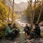 Four people sitting around a small outdoor stove fire at a rocky riverside campsite, with mountains, trees, and tents in the background.
