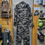 Camouflage jumpsuit displayed on a hanger against a wooden wall with outdoor gear.