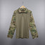 A military-style pullover shirt with a solid tan body and camouflage sleeves, hanging on a wooden hanger against a diamond plate metal door.