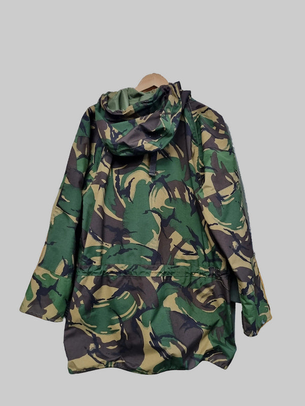 A hooded camouflage jacket with a green, black, and tan pattern, hanging on a wooden hanger against a solid gray background.