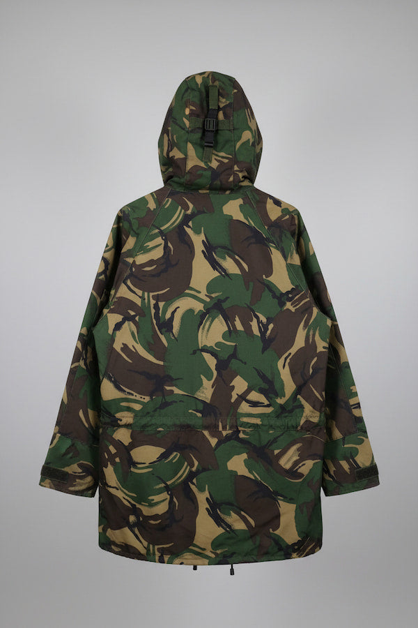 A hooded camouflage jacket shown from the back, featuring an all-over abstract leaf-like pattern in green, brown, and tan, photographed against a neutral gray background.