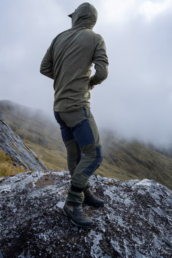 A person standing on a rocky mountain ridge, viewed from behind, wearing an olive green hooded jacket, dark blue and gray technical pants, and hiking boots, with misty hills in the background.