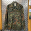 Camouflage military jacket with German flag patches on a hanger against a metallic wall.