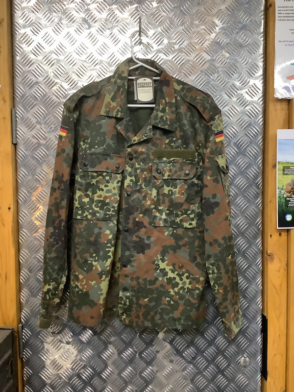Camouflage military jacket with German flag patches on a hanger against a metallic wall.