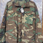 A woodland camouflage military-style jacket with four front flap pockets, hanging on a black hanger against a silver diamond plate metal background. A white hangtag is attached near the collar.