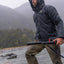 Man in a raincoat holding a rifle by a river with mountains in the background