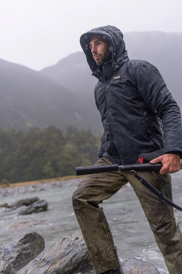 Man in a raincoat holding a rifle by a river with mountains in the background