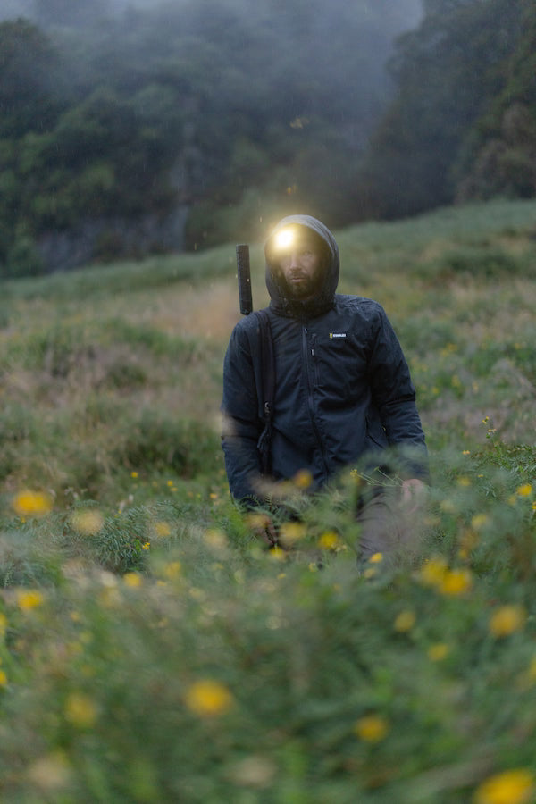 Person in a dark raincoat with a headlamp standing in a field with yellow flowers and greenery.