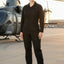 A woman stands on an airfield tarmac wearing a black one-piece jumpsuit and black boots, with a military helicopter blurred in the background.