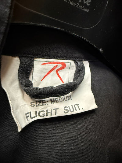 Close-up of a black flight suit interior label with a white tag reading 'SIZE: MEDIUM FLIGHT SUIT' and a red stylized logo, plus partial text 'of New Zealand' at the top.