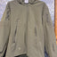 An olive green hooded softshell jacket with multiple zippered pockets, hanging on a black hanger against a textured diamond plate metal background.