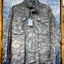 A military-style field jacket in a digital camouflage pattern, hanging on a black hanger against a textured diamond plate metal background.