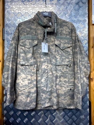 A military-style field jacket in a digital camouflage pattern, hanging on a black hanger against a textured diamond plate metal background.
