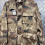A military-style field jacket in a digital camouflage pattern, hanging on a wooden hanger against a textured diamond plate metal background. The jacket features multiple flap pockets and a high collar.