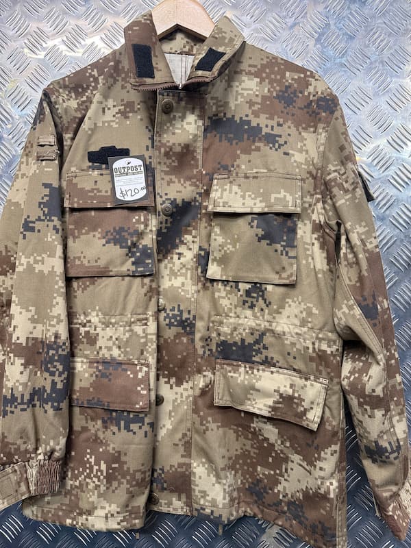 A military-style field jacket in a digital camouflage pattern, hanging on a wooden hanger against a textured diamond plate metal background. The jacket features multiple flap pockets and a high collar.