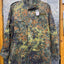 A long-sleeve military-style camouflage jacket hanging on a hanger against a diamond plate metal door. The jacket features a German Flecktarn camouflage pattern with green, brown, black, and tan spots, and has a visible hangtag.