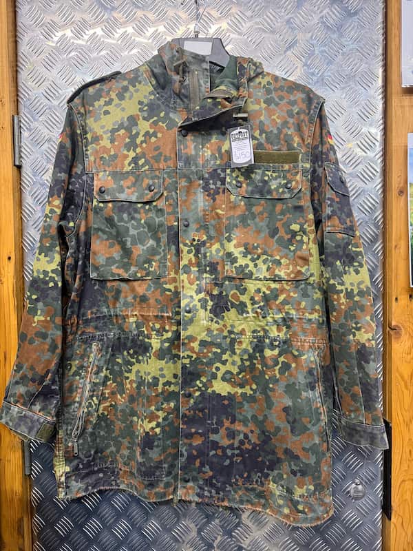 A long-sleeve military-style camouflage jacket hanging on a hanger against a diamond plate metal door. The jacket features a German Flecktarn camouflage pattern with green, brown, black, and tan spots, and has a visible hangtag.