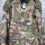 A military-style woodland camouflage jacket with green, brown, and tan patterns, featuring a high collar, shoulder epaulets, and four front flap pockets, displayed on a hanger against a diamond plate background.