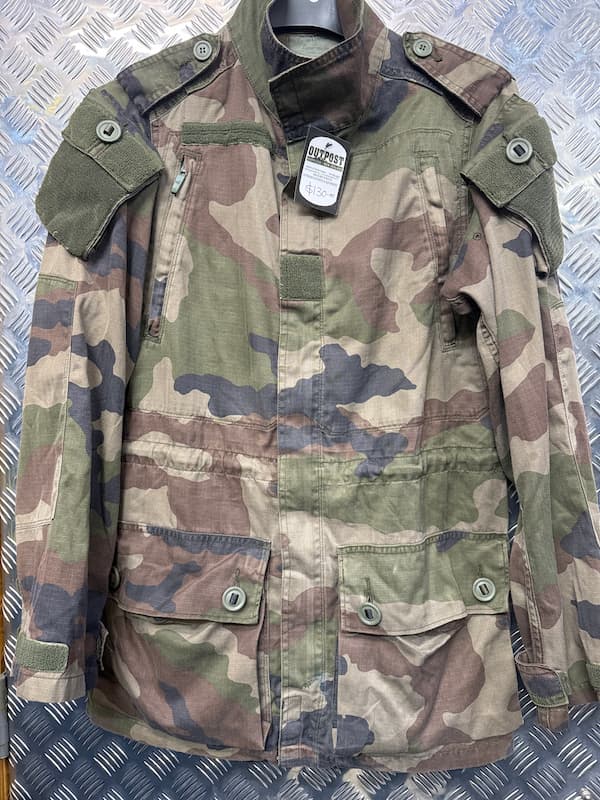 A military-style woodland camouflage jacket with green, brown, and tan patterns, featuring a high collar, shoulder epaulets, and four front flap pockets, displayed on a hanger against a diamond plate background.