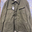 An olive green military-style utility jacket hanging on a wooden hanger against a blue diamond-plate metal background. The jacket features a button-up front, a pointed collar, and four front flap pockets.