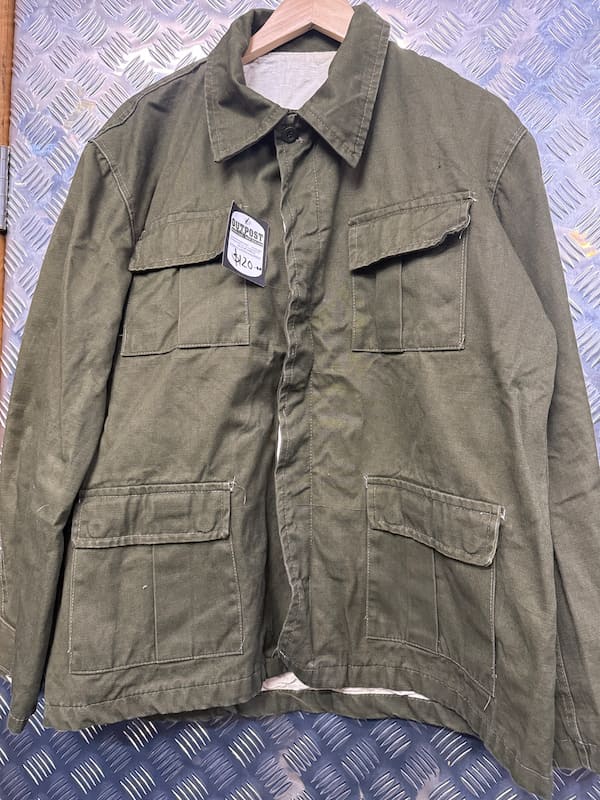 An olive green military-style utility jacket hanging on a wooden hanger against a blue diamond-plate metal background. The jacket features a button-up front, a pointed collar, and four front flap pockets.