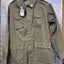An olive green military-style field jacket with multiple front pockets and shoulder epaulets, hanging on a dark hanger against a blue diamond-plate metal background.