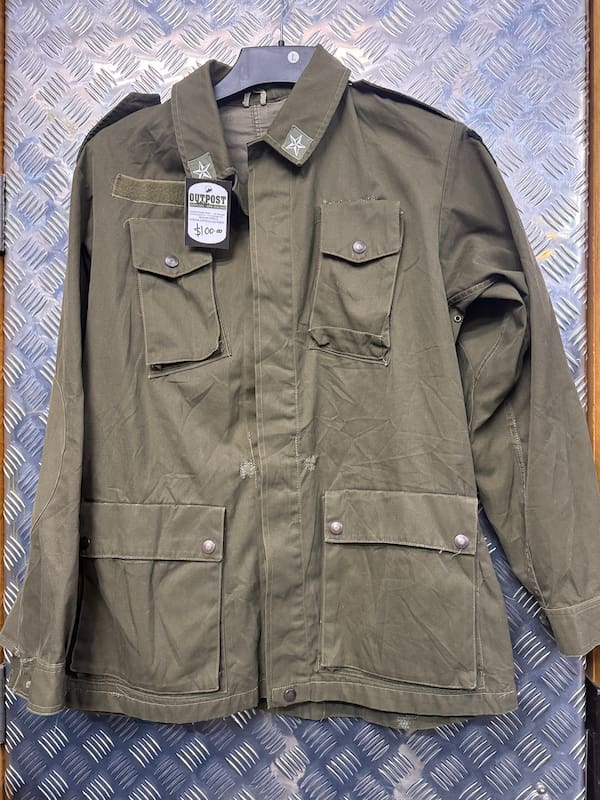 An olive green military-style field jacket with multiple front pockets and shoulder epaulets, hanging on a dark hanger against a blue diamond-plate metal background.