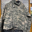A digital camouflage utility jacket in a gray and beige pattern, hanging on a black hanger against a diamond plate metal wall.