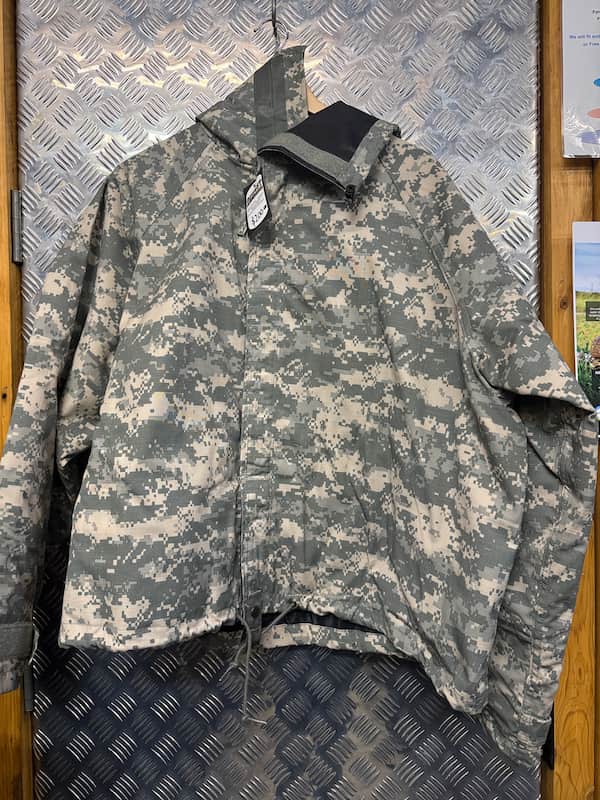 A digital camouflage utility jacket in a gray and beige pattern, hanging on a black hanger against a diamond plate metal wall.