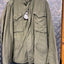 An olive green military-style field jacket with a high collar, button front, and multiple flap pockets, hanging against a diamond plate metal background.