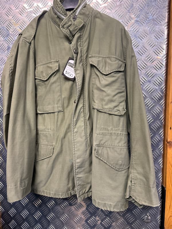An olive green military-style field jacket with a high collar, button front, and multiple flap pockets, hanging against a diamond plate metal background.