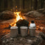 Three camping mugs and a metal water bottle arranged on a rock beside a campfire in a forest setting.