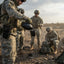 Soldiers in digital camouflage uniforms preparing water in an outdoor desert setting, with one soldier pouring from a black canteen into a cup.