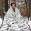 A person wrapped in a reflective silver emergency blanket or poncho, standing outdoors in a snowy wooded area.