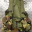 An olive green tactical vest with camouflage utility pouches on the sides, hanging on a metal hanger over a diamond plate metal background.