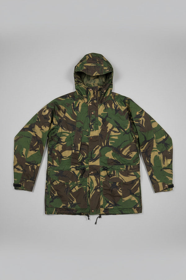 A hooded camouflage jacket in green, brown, and tan tones, laid flat against a neutral gray background.