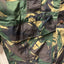 Close-up of a camouflage jacket or vest showing green, brown, and tan camo pattern with a flap pocket and drawstring.