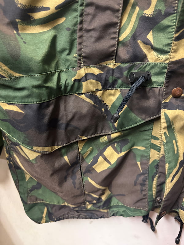 Close-up of a camouflage jacket or vest showing green, brown, and tan camo pattern with a flap pocket and drawstring.