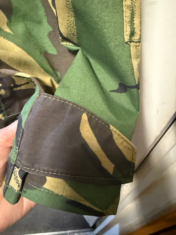 Close-up of a camouflage-patterned garment sleeve with a dark brown cuff tab, held by a hand against an indoor background.