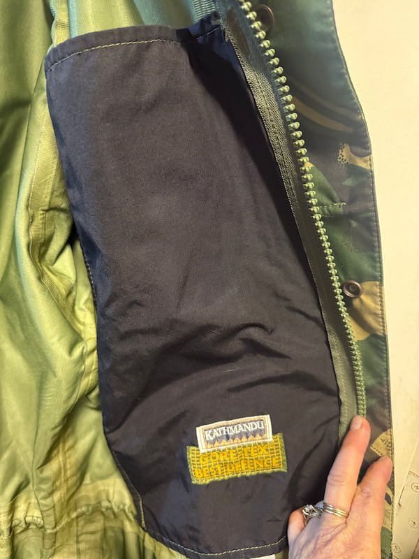 Close-up of a Katmandu jacket interior showing a dark navy inner pocket with a visible Katmandu brand patch and a yellow hook-and-loop section, next to an olive green outer fabric and zipper.