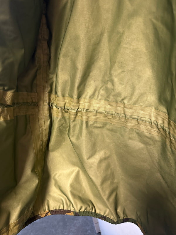 Close-up of a shiny olive green waterproof jacket or rain cover showing the fabric texture, horizontal seam stitching, and elasticized bottom edge.