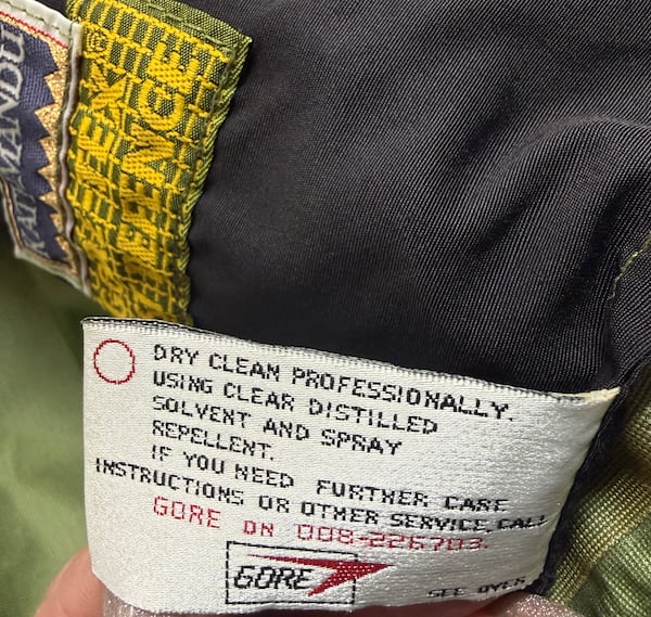 Close-up of a garment's interior care label showing the Gore brand and care instructions, with a dark gray fabric body and a yellow patterned trim.