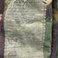 Close-up of a camouflage garment interior label showing printed text including 'Height/Chest 190/120' and care symbols, with a hand holding the fabric edge.